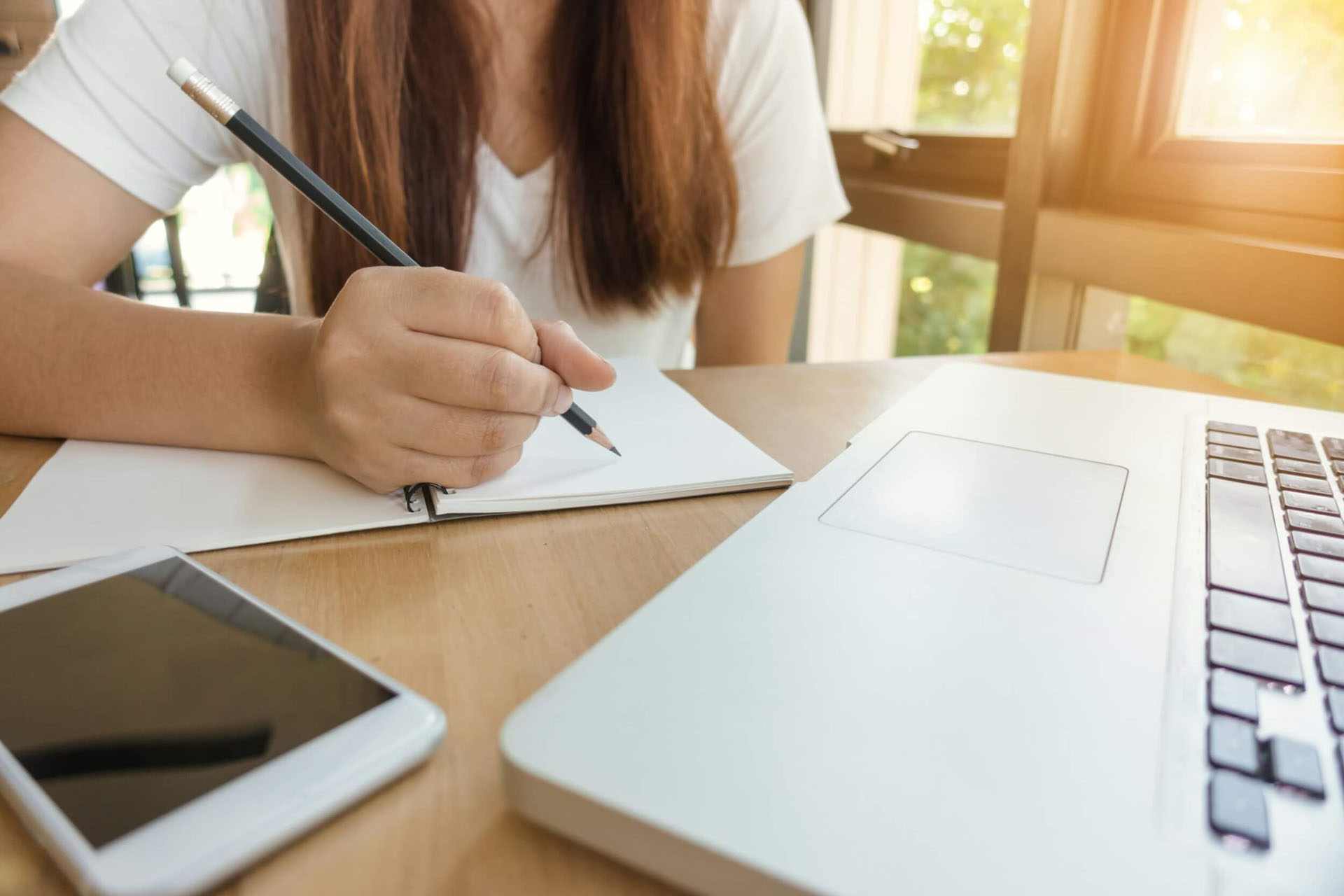 A girl sitting at a desk beside an window with a notepad, laptop, and phone