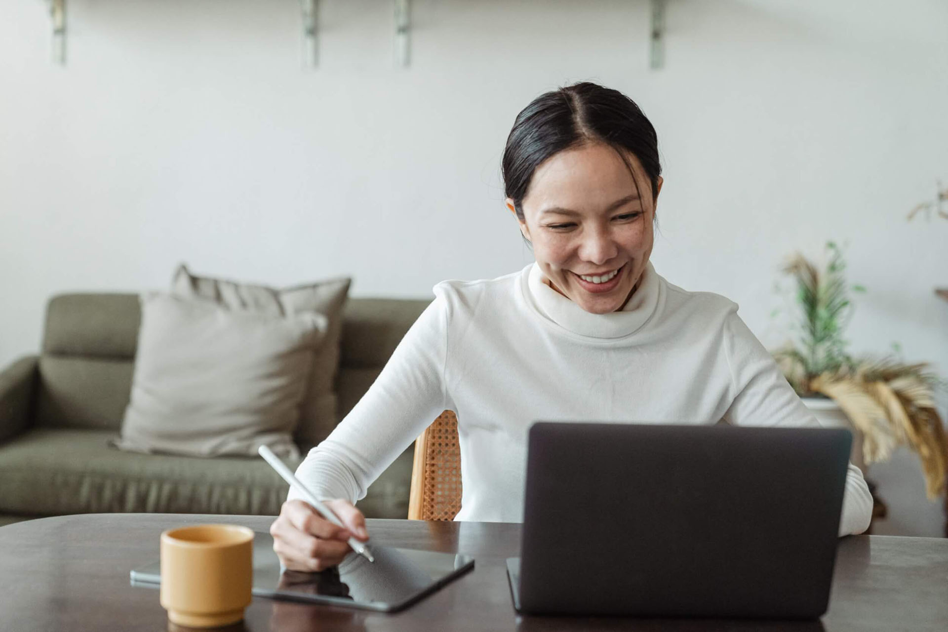 A woman sitting in a living room, smiling at a laptop, working on a tablet.