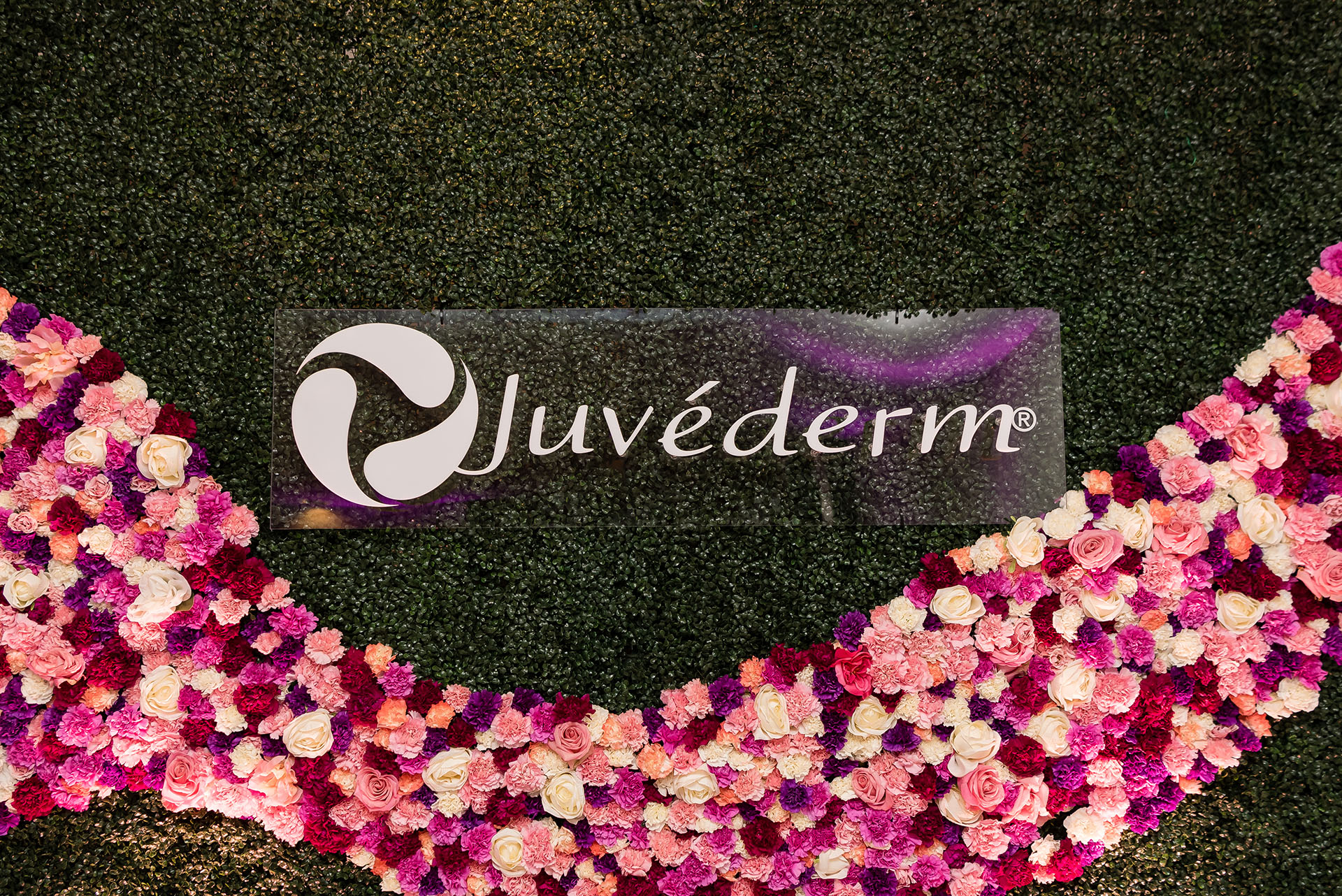 A large clear sign with the words Juvederm hanging on a floral and plant wall