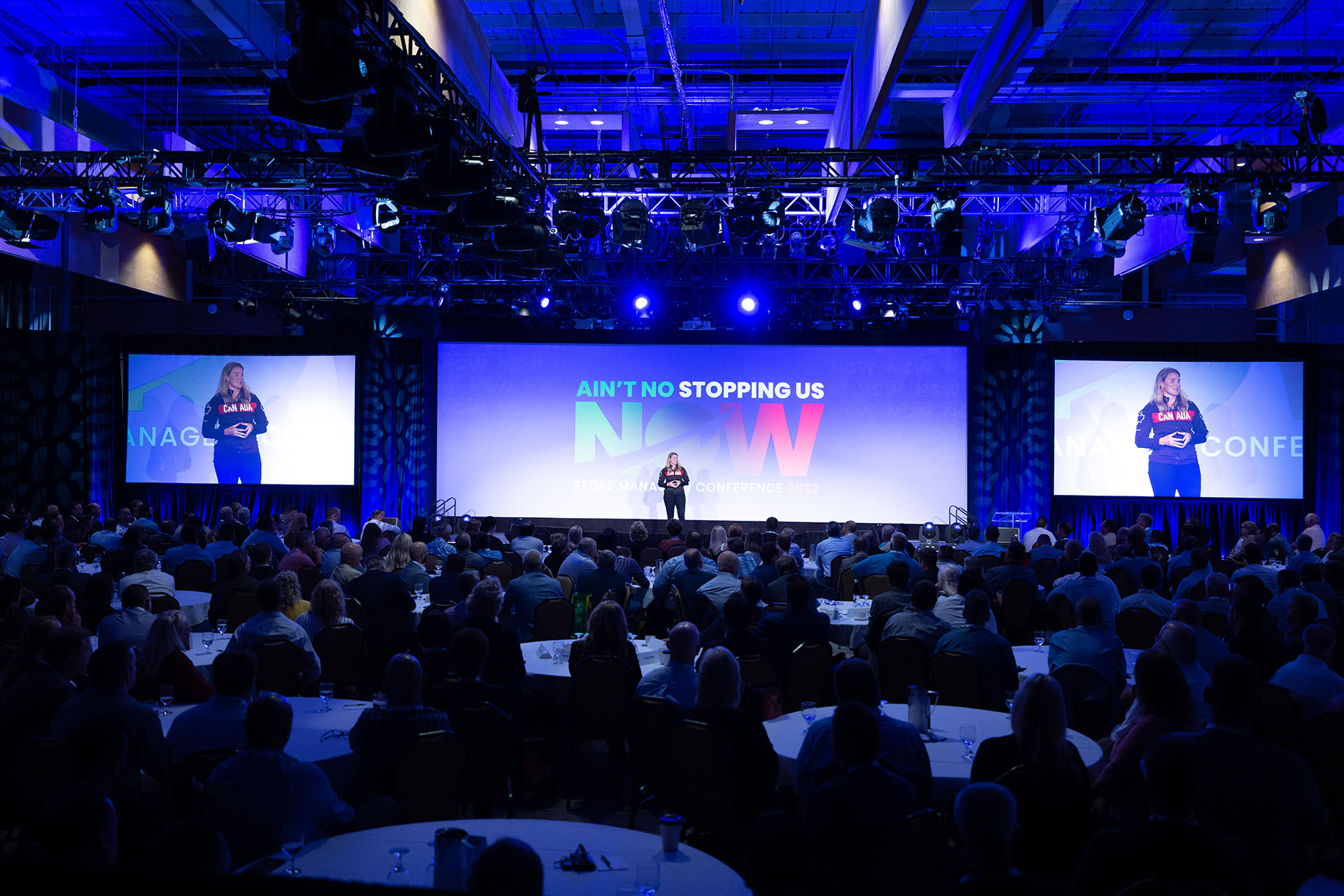 A woman on stage presenting at a conference in front of a group of people sitting at tables