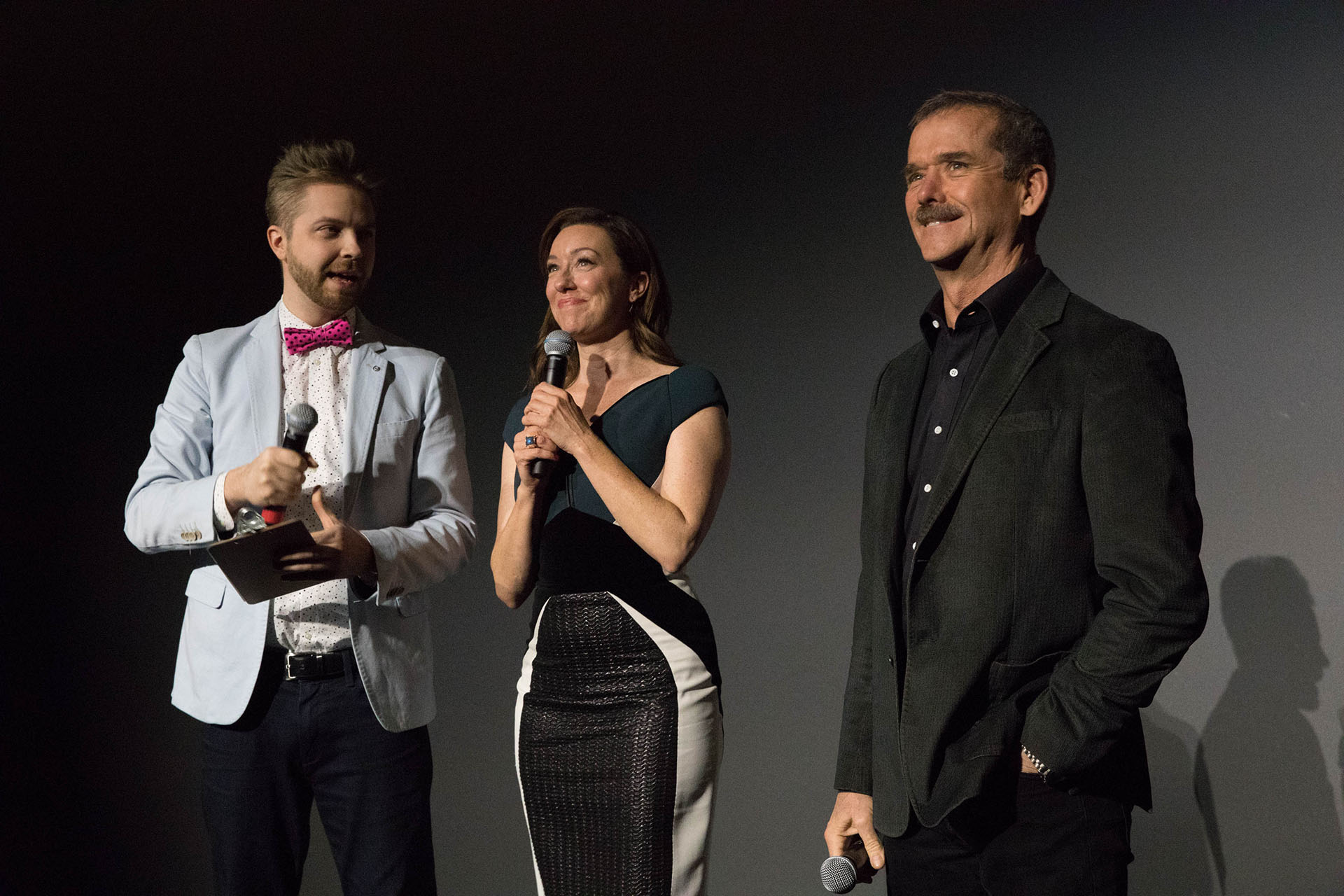 two men and one woman standing with microphones in front of a black wall