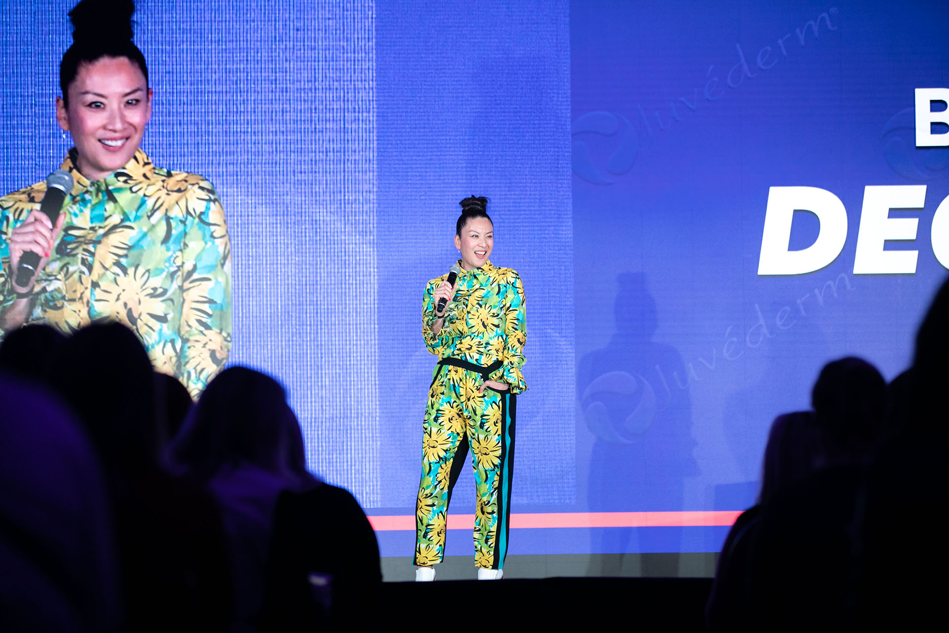 A woman standing on a stage in front of a large screen wearing a floral print suit