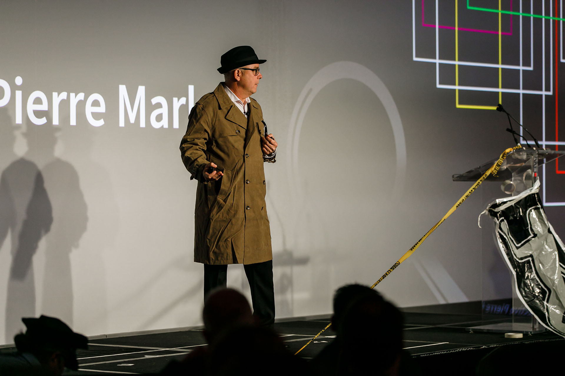 A man in a long trench coat and hat standing on a stage in front of a crowd